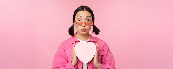 심장 모양의 상자를 선물로 내보이는 귀여운 아시아인 소녀, 놀란 모습 과 흥분된 모습, 선글라스를 쓰고 핑크 색 배경 위에 서 있는 낭만적 인 현재의 개념 — 스톡 사진