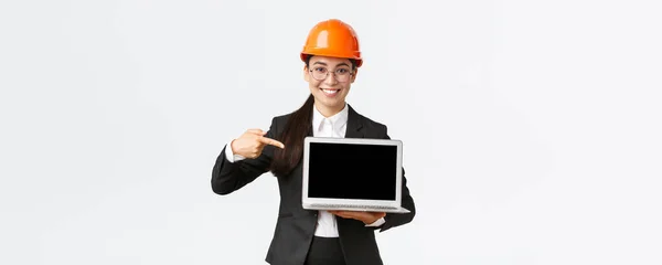 Sonriendo complacido asiático ingeniero jefe mostrando gráfico con beneficio empresarial, haciendo presentación durante la reunión de inversores, usando casco de seguridad, apuntando a la pantalla del ordenador portátil, fondo blanco — Foto de Stock