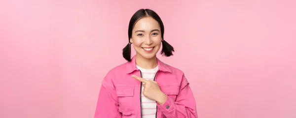 Portrait de belle fille asiatique souriante, doigt pointant vers la gauche, montrant la publicité, bannière promo, debout sur fond rose — Photo