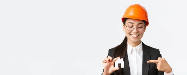 웃고 있는 이 시안 여성 기술자, 헬멧을 쓰고 정장을 입은 건축가, 집에 미니어처를 들고 작은 집을 가리키며, 건물짓는 일을 하고 있는 백인 배경의 건축가 — 스톡 사진