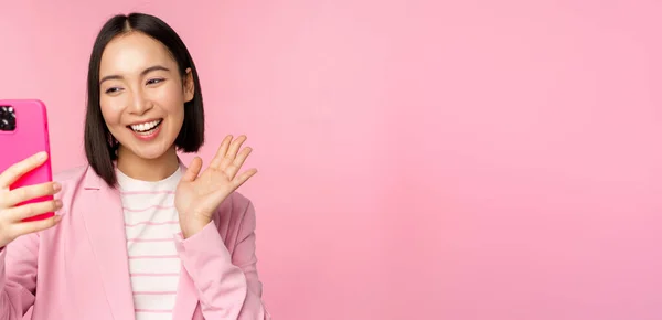 Stylowe azjatyckie businesswoman, dziewczyna w garniturze robi selfie na smartfonie, wideo czat z aplikacją na telefon komórkowy, pozowanie na różowym tle studio — Zdjęcie stockowe