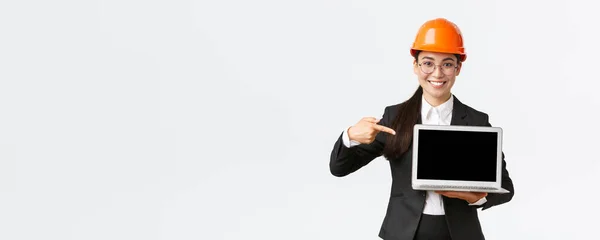 Sonriendo complacido asiático ingeniero jefe mostrando gráfico con beneficio empresarial, haciendo presentación durante la reunión de inversores, usando casco de seguridad, apuntando a la pantalla del ordenador portátil, fondo blanco — Foto de Stock