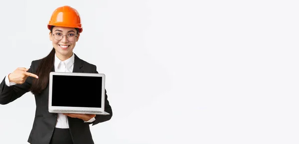 Χαμογελώντας ευχαριστημένος ασιατική επικεφαλής μηχανικός δείχνει διάγραμμα με επιχειρηματικό κέρδος, κάνοντας παρουσίαση κατά τη διάρκεια της συνάντησης των επενδυτών, φορώντας κράνος ασφαλείας, δείχνοντας στην οθόνη του φορητού υπολογιστή, λευκό φόντο — Φωτογραφία Αρχείου