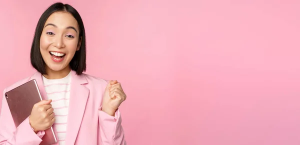Захоплена азіатська бізнес-леді з цифровим планшетом, кричить з радістю, тріумфує, стоїть над рожевим тлом у костюмі — стокове фото