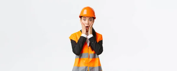 Sorprendido y preocupado asiático mujer ingeniero tener problema en la construcción de área, mirar el proyecto asombrado con pánico cara, cogido de la mano cerca de la boca y jadeando preocupado, de pie fondo blanco — Foto de Stock