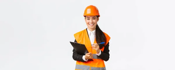 Confiante sorrindo mulher asiática engenheiro de construção, mulher industrial em segurança capacete visita área de construção para inspeção, anotando notas na área de transferência e olhando satisfeito, fundo branco — Fotografia de Stock