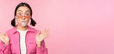 Havalı Asyalı kız sakız şişiriyor, sakız çiğniyor, güneş gözlüğü takıyor, pembe arka planda poz veriyor.