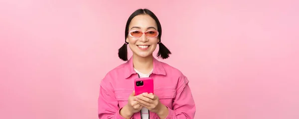 Portrait de fille coréenne dans des lunettes de soleil élégantes tenant le téléphone mobile, en utilisant l'application smartphone, debout sur fond rose — Photo