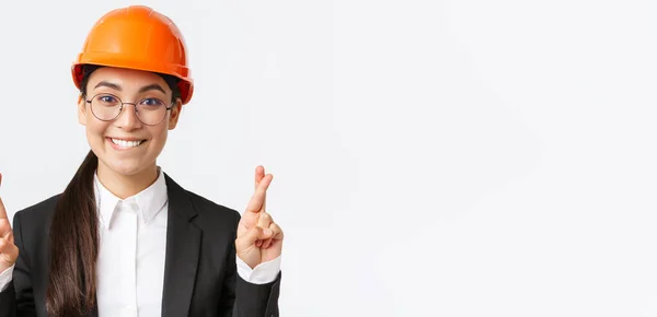 Primer plano de emocionado esperanzado asiático ingeniero, gerente de construcción en casco de seguridad y traje de negocios suplicando por el gusto, dedos cruzados buena suerte y morder el labio tentador, fondo blanco — Foto de Stock