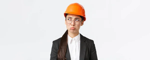 Primer plano de sombrío molesto asiático mujer ingeniero en casco de seguridad y traje de negocios quejándose, buscando decepcionado, lloriqueando con cara de enfurruñamiento, trabajo involuntario los fines de semana, mirando a la izquierda molestado — Foto de Stock