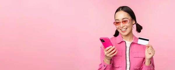Интернет-магазины. Улыбающаяся азиатка-покупатель, держит смартфон и кредитку, платит мобильным приложением, стоит на розовом фоне — стоковое фото