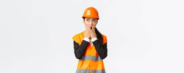 Geschokt en bezorgd aziatische vrouwelijke ingenieur, architect in veiligheidshelm en reflecterende jas, kijken linksboven hoek en hijgend bezorgd, staren verbaasd, staande witte achtergrond — Stockfoto