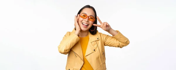 Retrato de chica moderna asiática con estilo, con gafas de sol y chaqueta amarilla, mostrando paz, gesto de signo v, de pie sobre fondo blanco, cara sonriente feliz —  Fotos de Stock