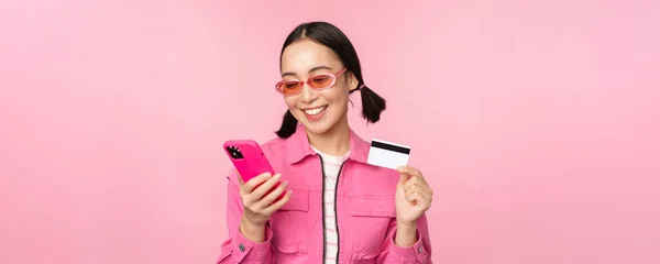 Compras en línea. Sonriendo chica asiática shopper, sosteniendo teléfono inteligente y tarjeta de crédito, pagando en aplicación móvil, de pie sobre fondo rosa —  Fotos de Stock