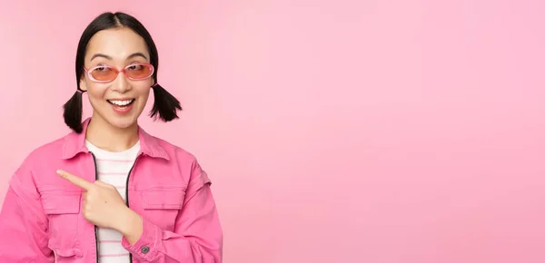 Portrait de fille asiatique souriante en tenue élégante, lunettes de soleil, doigt pointant vers la gauche, montrant la publicité, bannière, debout sur fond rose — Photo