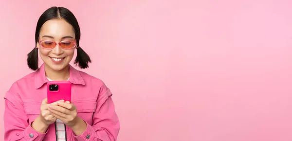 用智能手机刻画戴太阳镜的亚洲女孩。女人看手机，浏览应用程序，站在粉色背景上 — 图库照片