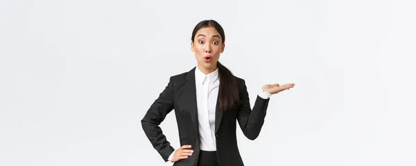Удивительная азиатская бизнесвумен в черном костюме указывает рукой вправо и смотрит в камеру интересуется, обсуждая объявление, показывая баннер, стоя на белом фоне заинтригован — стоковое фото