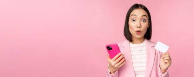 Harika bir iş kadını, takım elbiseli Asyalı bir kız kredi kartı ve cep telefonu gösteriyor, internetten sipariş veriyor, akıllı telefonla alışveriş yapıyor, pembe arka planda duruyor. Reklam kavramı