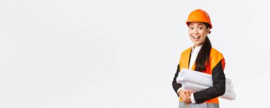 Şaşırmış ve heyecanlı Asyalı kadın mimar, güvenlik kaskı ve yansıtıcı ceket giymiş baş mühendis, neşeli ve mutlu görünüyor, etkilenmiş gülümsüyor, elinde binanın planlarını tutuyor.