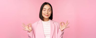 Sakin olun. Gülümseyen genç Asyalı kadın meditasyon yapıyor, yoga yapıyor, iş yerinde farkındalık ve gevşeme gösteriyor, nefes alıyor, keyifli bir şekilde havayı soluyor, pembe arka planda duruyor.