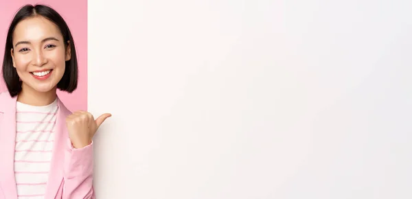 Πορτρέτο της χαμογελαστή ασιάτισσα επιχειρηματίας με κοστούμι, εταιρική κυρία δείχνοντας δάχτυλο σε λευκό άδειο τοίχο, πίνακα με πληροφορίες ή διαφήμιση, στέκεται πάνω από ροζ φόντο — Φωτογραφία Αρχείου