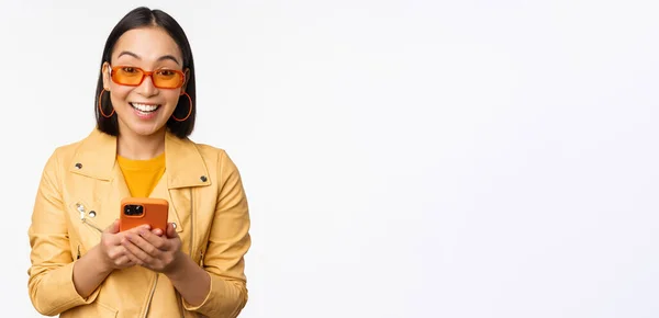Moderna ragazza asiatica in occhiali da sole utilizzando il suo telefono cellulare, sorridente e guardando felice, in posa su sfondo bianco — Foto Stock