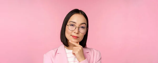 Koreanische Geschäftsfrau denkt, trägt eine Brille, blickt nachdenklich in die Kamera, trifft Entscheidungen, steht vor rosa Hintergrund — Stockfoto