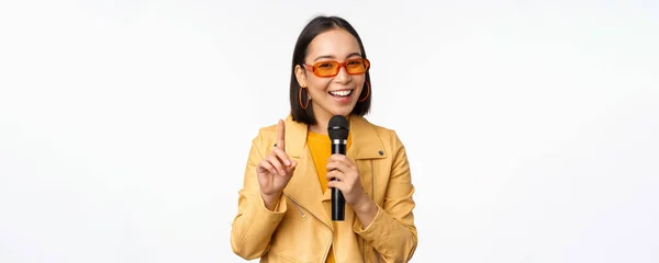 Retrato de hermosa mujer asiática en gafas de sol, chica elegante cantando, dando discurso con micrófono, sosteniendo micrófono y sonriendo, de pie sobre fondo blanco — Foto de Stock
