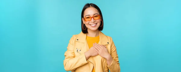 Portrait of asian woman smiling, holding hands on heart and looking with tenderness, care at camera, thankful emotion, standing over blue background — Stock Photo, Image