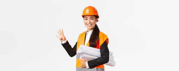 Mosolygó magabiztos ázsiai női mérnök, építőipari vezető a biztonsági sisak, szállít tervrajzokat, mutatja négy ujját, biztosítja az épület időben kész, fehér háttér vidám — Stock Fotó
