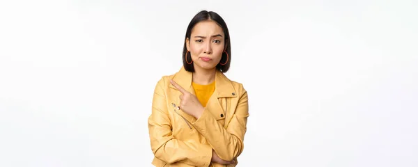 Портрет стильной азиатской девушки, смотрящей с сомнением, скептически настроенным лицом, указывающей пальцем на логотип или рекламу, стоящей на белом фоне — стоковое фото