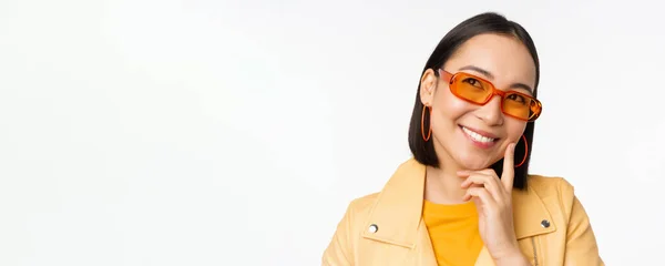Close up retrato de mulher asiática pensando, vestindo óculos de sol e sorrindo, olhando para cima pensativo, de pé sobre fundo estúdio branco — Fotografia de Stock