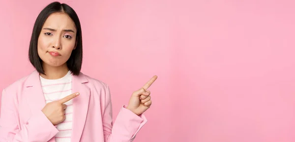 Scettica donna d'affari asiatica, commessa imbronciata e guardando con disapprovazione mentre indicava le dita a destra, mostrando cattive informazioni, notizie sconvolgenti, in piedi su sfondo rosa — Foto Stock