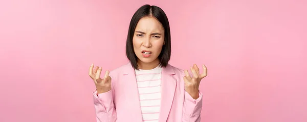 Ritratto di donna asiatica arrabbiata in giacca e cravatta, pugni stretti e sguardo furioso, indignato di smth male, in piedi su sfondo rosa — Foto Stock