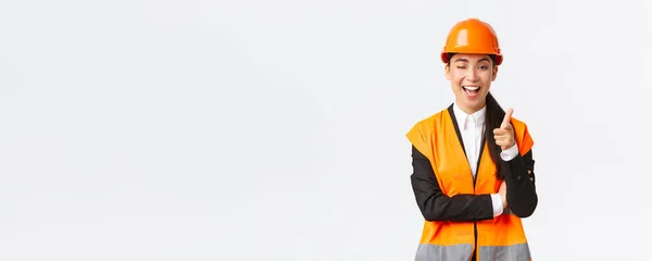 穿着安全帽和反光夹克，微笑专业的亚洲女施工经理，招揽新大楼的入住、眨眼和对着相机指指点点，向客户展示最佳地产 — 图库照片