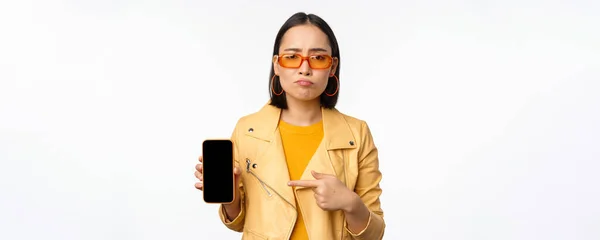 Ritratto di donna asiatica triste in occhiali da sole, puntando il dito verso l'interfaccia dell'app del telefono cellulare, mostrando l'applicazione dello smartphone, in piedi su sfondo bianco — Foto Stock