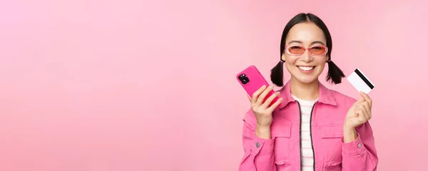 Vacker koreansk kvinna som håller smartphone, kreditkort, ler mot kameran, köper online, handlar med mobiltelefon, står över rosa bakgrund — Stockfoto