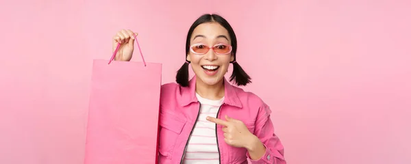 买东西戴着太阳镜的时髦亚洲女孩，带着购物袋和微笑，推荐商店里的促销活动，站在粉色背景上 — 图库照片