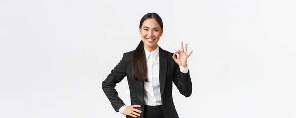 Професійна жінка-менеджер в чорному костюмі виглядає наполегливо, заохочує все добре, гарантує роботу, показує нормальний жест і посміхається в схваленні, стоячи задоволені на білому тлі — стокове фото