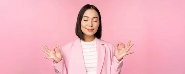 Stai calmo. Sorridente giovane donna asiatica meditare, praticare yoga, consapevolezza e relax sul lavoro, respirare, inalare aria con il viso soddisfatto, in piedi su sfondo rosa — Foto Stock