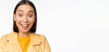 Etkilenmiş Asyalı kızı yakından çek, kameraya hayret içinde bak, beyaz arka plan üzerinde günlük giysiler içinde dur.