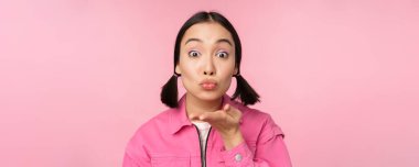 Güzel, aptal Asyalı bir kızın yakın portresi, hava öpücüğü, kameraya mwah, üfleme, pembe arka plan üzerinde durmak