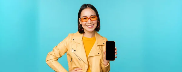 Bild av ung snygg asiatisk kvinnlig modell i trendiga solglasögon, visar mobiltelefon skärm, ansökan på smartphone gränssnitt, står över blå bakgrund — Stockfoto