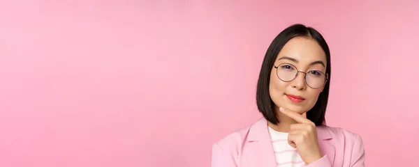 Donna d'affari coreana pensando, indossando gli occhiali, guardando pensieroso alla fotocamera, prendere decisioni, in piedi su sfondo rosa — Foto Stock