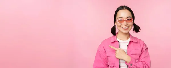 Retrato de sorrindo ásia menina no elegante roupa, óculos de sol, apontando dedo esquerdo, mostrando anúncio, banner, pé sobre rosa fundo — Fotografia de Stock