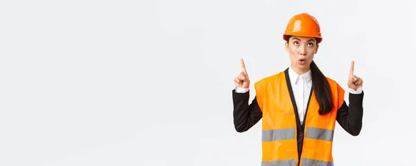 Construção, construção e conceito industrial. Surpreendido e curioso asiático feminino gerente de construção, engenheiro em capacete de segurança e roupas reflexivas, apontando os dedos para cima em banner interessante — Fotografia de Stock