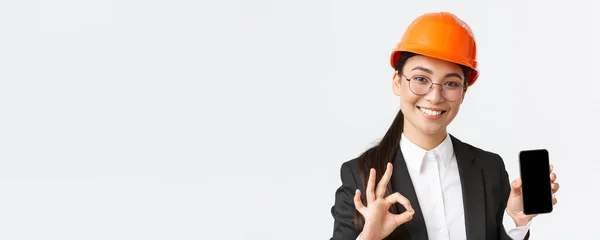 Nöjd ung kvinnlig asiatisk ingenjör, arkitekt i kostym och säkerhetshjälm visar mobiltelefon skärm och göra okej gest, godkänna, rekommendera ansökan, vit bakgrund — Stockfoto