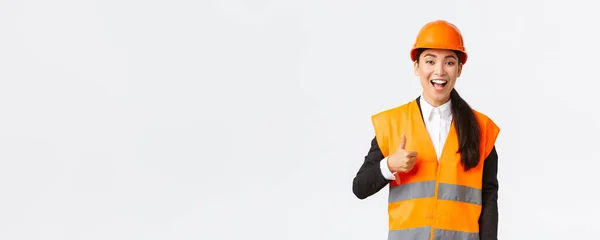 Apoyo y complacido feliz asiática jefe ingeniero en casco de seguridad mostrando pulgares hacia arriba en la aprobación, satisfecho con los trabajos de construcción. Mujer industrial en zona de inspección de chaqueta reflecive —  Fotos de Stock