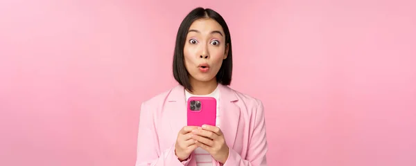 Retrato de mulher de negócios asiática com rosto surpreso, usando aplicativo smartphone, vestindo terno de negócios. Menina coreana com telefone celular e expressão facial animado, fundo rosa — Fotografia de Stock