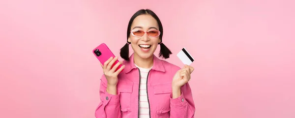 Nowoczesna piękna azjatycka dziewczyna, śmiech i uśmiech z telefonu komórkowego, karta kredytowa, zakupy online, płacenie smartfonem, stojąc na różowym tle — Zdjęcie stockowe